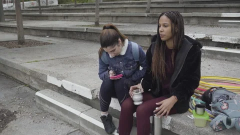 Two women drinking mate cup infusion seated in urban park 库存影片 130431845