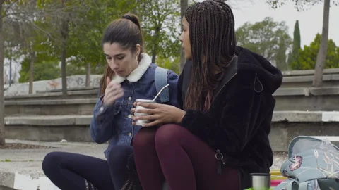 Two women drinking mate cup infusion seated in urban park 库存影片 130432409