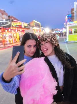 Two women joking while taking selfie holding a cotton candy in a night fair Stock Photos