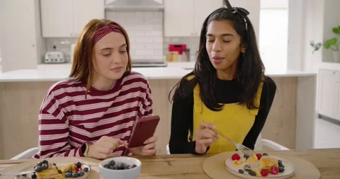 Two women at kitchen table, left tapping red phone showing pics, right eating Stock Footage 342607977