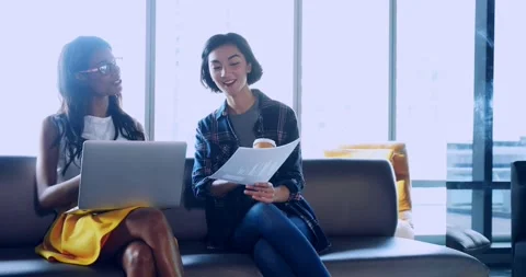 Two women in lounge, right leaning with papers, reviewing laptop, lifting cup, Stock Footage 330355253