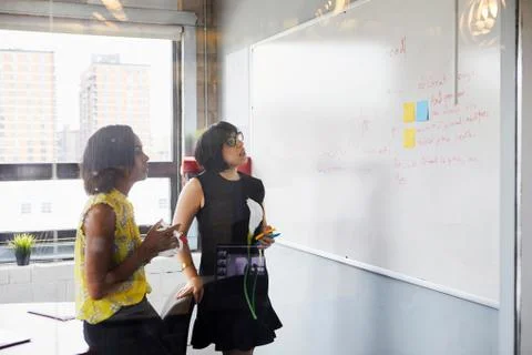 Two women in office, solving problem, using whiteboard, sticky notes stuck on Foto stock