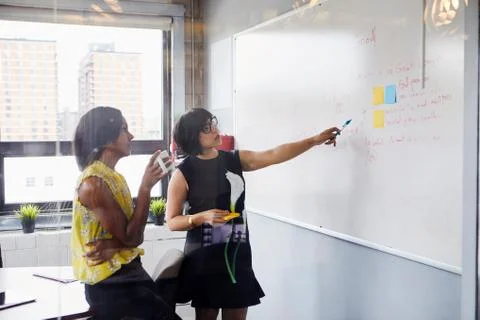 Two women in office, solving problem, using whiteboard, sticky notes stuck on Stock Photos