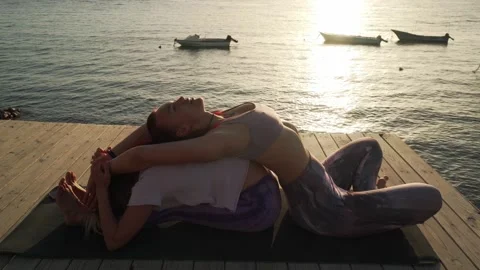 Two women practice seated forward and back bends yoga posture at seaside Stock Footage 130779312