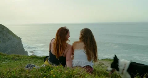 Two women sit side by side on a grassy hill overlooking the vast ocean. Stock Footage 334173147