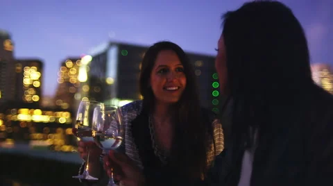 Two women sit at a table and have a glass of wine at a rooftop bar at sunset Video stock 44180367