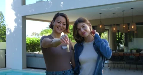 Two women standing poolside patio, presenting key toward camera, celebrating new Stock Footage 334999930