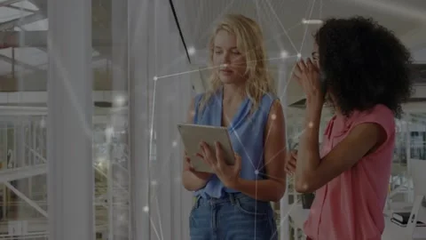 Two women tapping tablet, causing glowing node overlay across glass office, Video stock 330959540