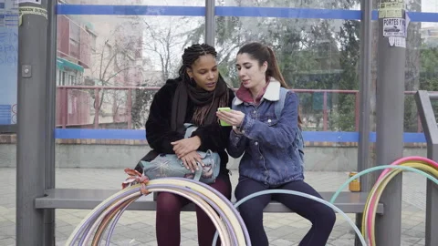 Two women using smartphone seated on bus stop 库存影片 130396865
