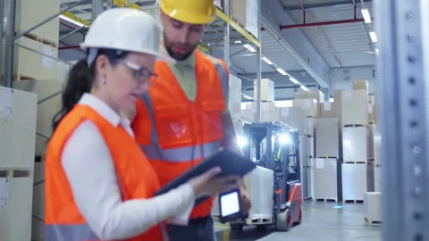 Two workers in hard hats and vests inspect cargo with a tablet in a warehouse. 库存影片 307998557