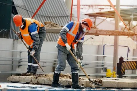 Two workers in hard hats, work clothes and a medical mask work with shovels at a Stock Photos