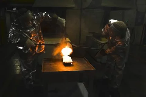 Two workers in heat-protective suits pour molten gold from a crucible into an Stock Photos