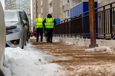 Two workers in yellow reflective vests sprinkle sand on a walkway from a cart. Stock Photos