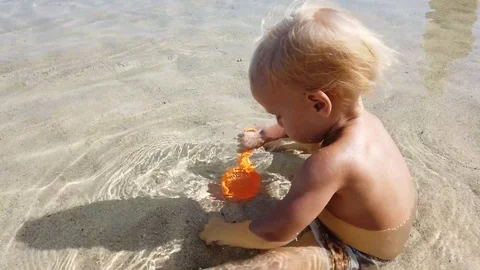 A two-year-old blond boy sits in clean sea water and plays with children's toys. Stock Footage 108577789