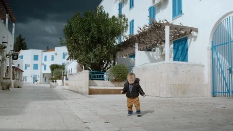 Two-year-old boy in a black jacket walks an empty street in on a sunny day Stock Footage 127079371