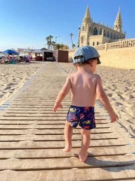 Two year old boy with cap and swimsuit walking along the boards of the beach Stock Photos
