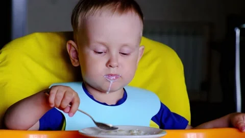 A two-year-old boy enjoys eating milk soup with a spoon, from a child's seat. Stock Footage 155369997