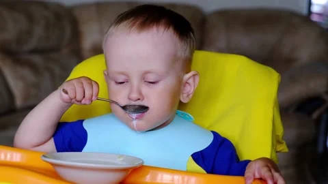 A two-year-old boy enjoys eating milk soup with a spoon, from a child's seat. Stock Footage 157105730