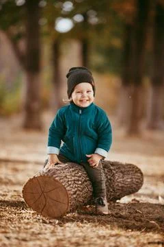 A two-year-old boy in a hat and jacket sits on a log in a Pine forest against Stock Photos
