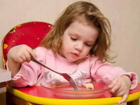 Two-year-old girl eats with a fork at table in kitchen Stock Photos