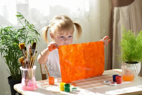 A two-year-old girl is sitting at a table and shows an orange sheet painted w Stock Photos