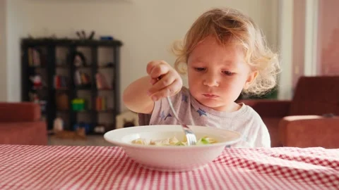 Two year old smiling baby boy sits at table in highchair and eats pasta with Vídeos de archivo 229479460