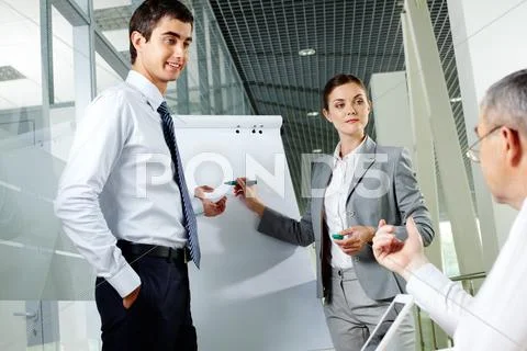 Two young associates by whiteboard looking at senior chief at meeting Stock Photos