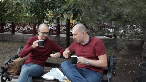 Two young bald guys at dinner are sitting in a park eating sandwiches and Stock Footage 95762764