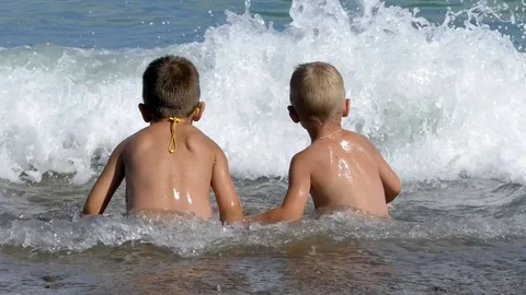 Two young boy, sitting together on a sandy shore bathed in a wave Stock Footage 73428090