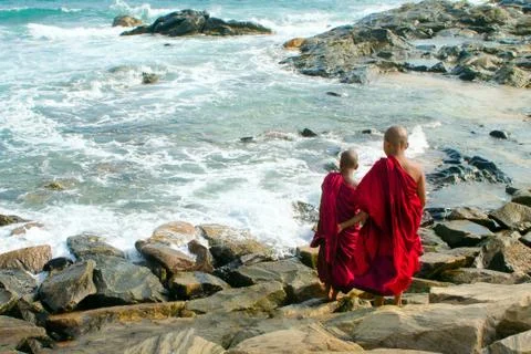 Two young Buddhist monks in red robes are standing at the shore of the ocean. Stock Photos