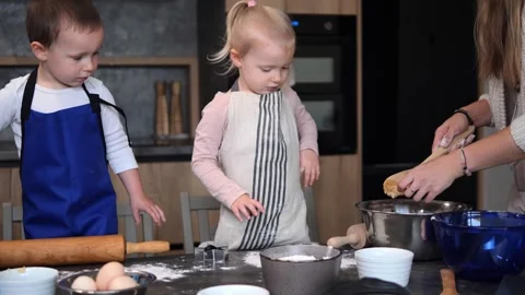 Two young children eagerly wait in a modern kitchen as their mother pours dough Stock Footage 289519090