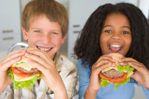 Two young children in kitchen eating cheeseburgers smiling Stock Photos