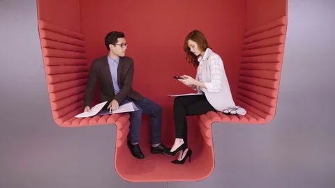 Two young coworkers talking about business tasks sitting in modern red zone for Stock Footage 80924168
