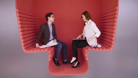 Two young coworkers talking about business tasks sitting in modern red zone for Stock Footage 80924674