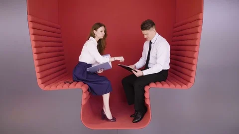 Two young coworkers talking about business tasks sitting in modern red zone for Stock Footage 80938674
