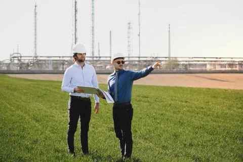 Two young engineers standing outdoors by oil refinery, discussing issues. Stock Photos