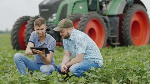 Two young farmers use a digital tablet in the field. Against tractor background Stock Footage 77427668