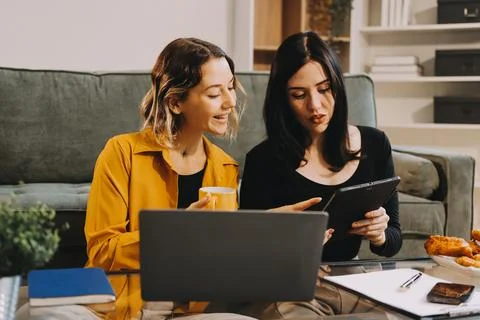 Two young female students using a laptop while sitting on a sofa at home. S.. Stock Photos
