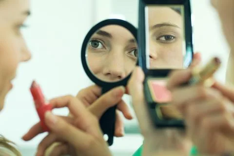 Two young friends looking at selves in hand mirrors, holding lipstick, cropped Stock Photos