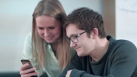 Two young friends sitting at table in school and working on smartphone 스톡 동영상 77508301