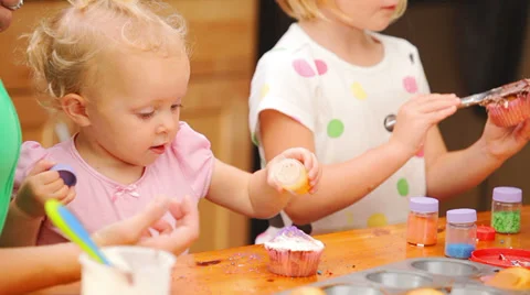 Two young girls help their mother put frosting and sprinkles on cupcakes Видео 33873017