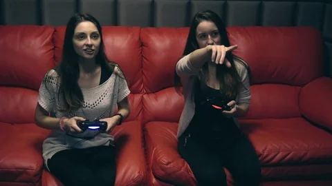 Two young girls playing video games with game controller on red sofa Stock Footage 83501367