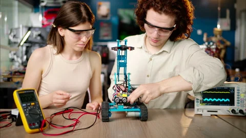 Two young happy engineers fixing a mechanical robot car in the workshop, using Stock Footage 247418208
