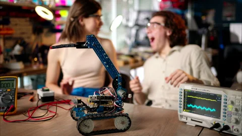Two young happy engineers fixing a mechanical robot car in the workshop, using Stock Footage 247418417