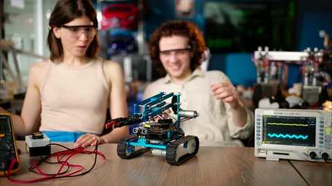 Two young happy engineers fixing a mechanical robot car in the workshop, using Stock Footage 247418740
