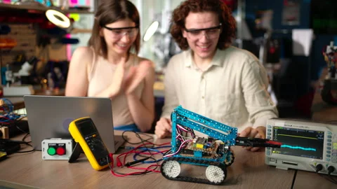 Two young happy engineers fixing a mechanical robot car in the workshop, Stock Footage 247419629