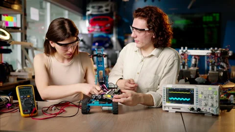 Two young happy engineers fixing a mechanical robot car in the workshop, using Stock Footage 247474888