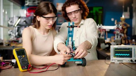 Two young happy engineers fixing a mechanical robot car in the workshop, using Stock Footage 247475258