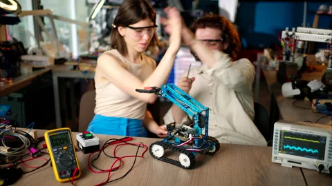 Two young happy engineers fixing a mechanical robot car in the workshop, using Stock Footage 247486162