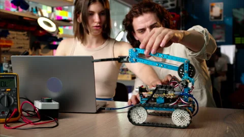 Two young happy engineers fixing a mechanical robot car in the workshop, Stock Footage 247486617